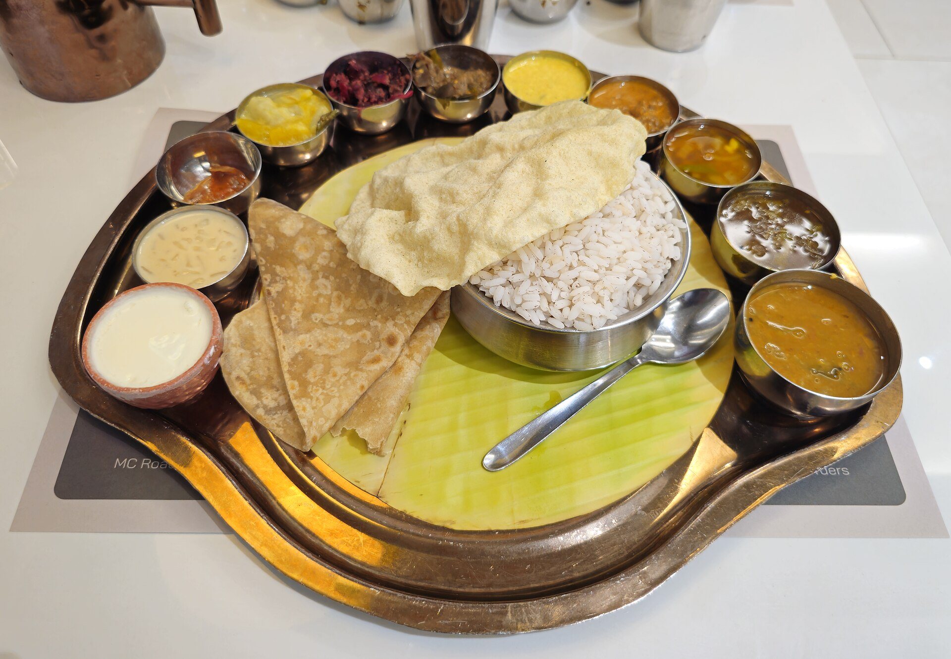 A brass thali laid on a banana leaf with fourteen separate components around a bowl of red rice — dals, curries, pickles, curd, buttermilk, a papad and wheat parathas